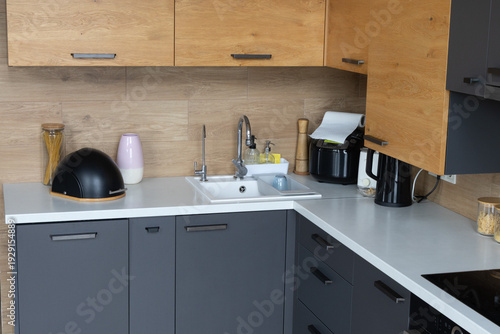 Modern L-shaped kitchen interior with a mix of oak wood upper cabinets and dark grey lower units. The scene features various kitchen accessories against a light wood-textured backsplash. 