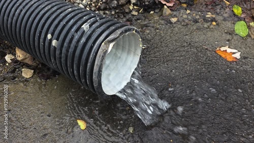Drain pipe pouring rainwater onto wet asphalt during an autumn storm, with colorful leaves fallen on the ground, representing a functional drainage system and water management