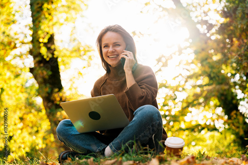 Young woman using laptop and talking on phone outdoors in fall park