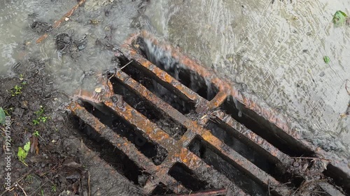 Heavy rainfall water runoff flowing into a rusty metal storm drain grate on a city street, showcasing urban drainage infrastructure during a downpour and flood prevention