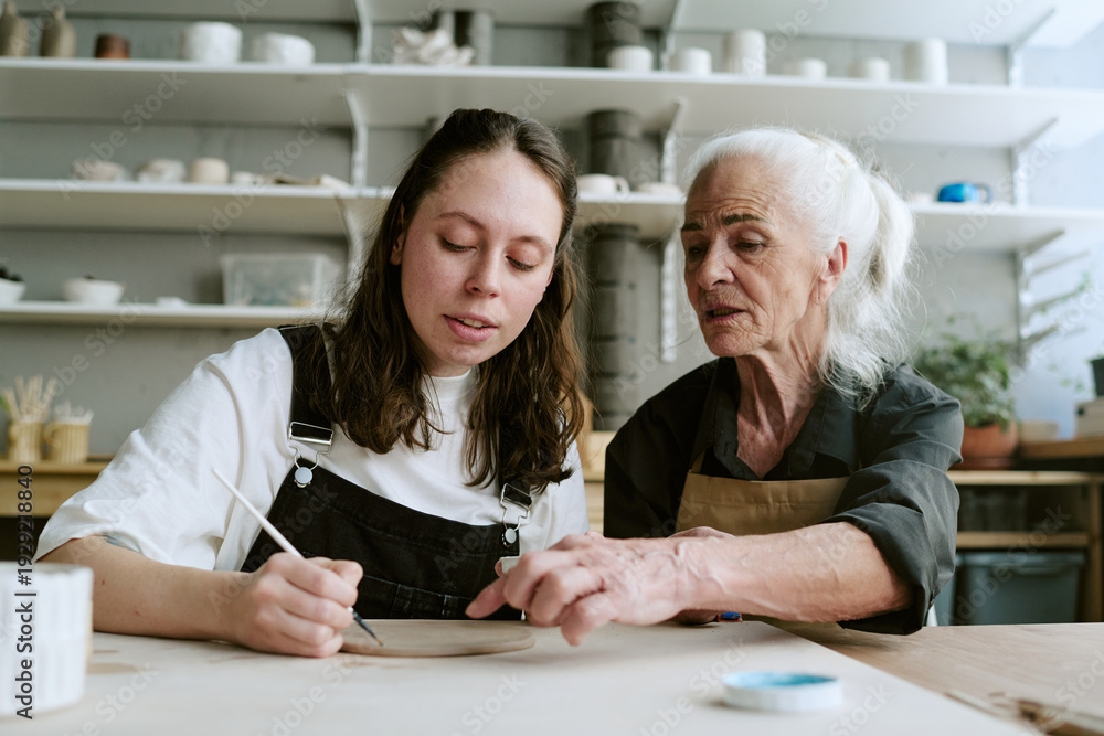 Obraz premium Caucasian young adult woman painting ceramic plate while senior Caucasian woman guiding her, both sitting at table in pottery studio, shelves with pottery visible in background