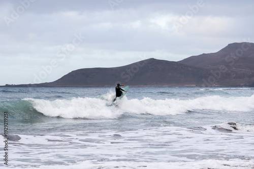 Surfer riding a breaking wave under an overcast sky, with dramatic volcanic mountains in the background.