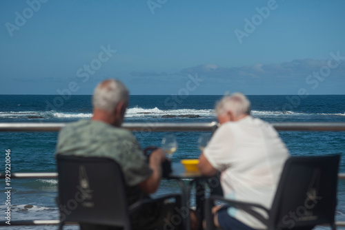 Senior couple seating by a seaside promenade railing, looking out at the ocean on a sunny day