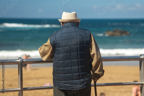 Senior man standing by a seaside promenade railing, looking out at the ocean on a sunny day