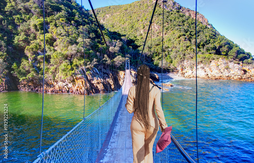 A beautiful young South African girl with braided hair watches the bridge - The suspension bridge over the storms river mouth within Tsitsikamma National Park - Eastern Cape in South Africa