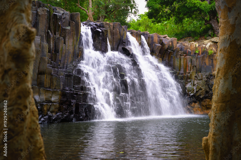 Fototapeta premium Rochester Falls in Mauritius flowing over distinctive basalt rock columns into a calm pool, surrounded by dense tropical forest and lush greenery.