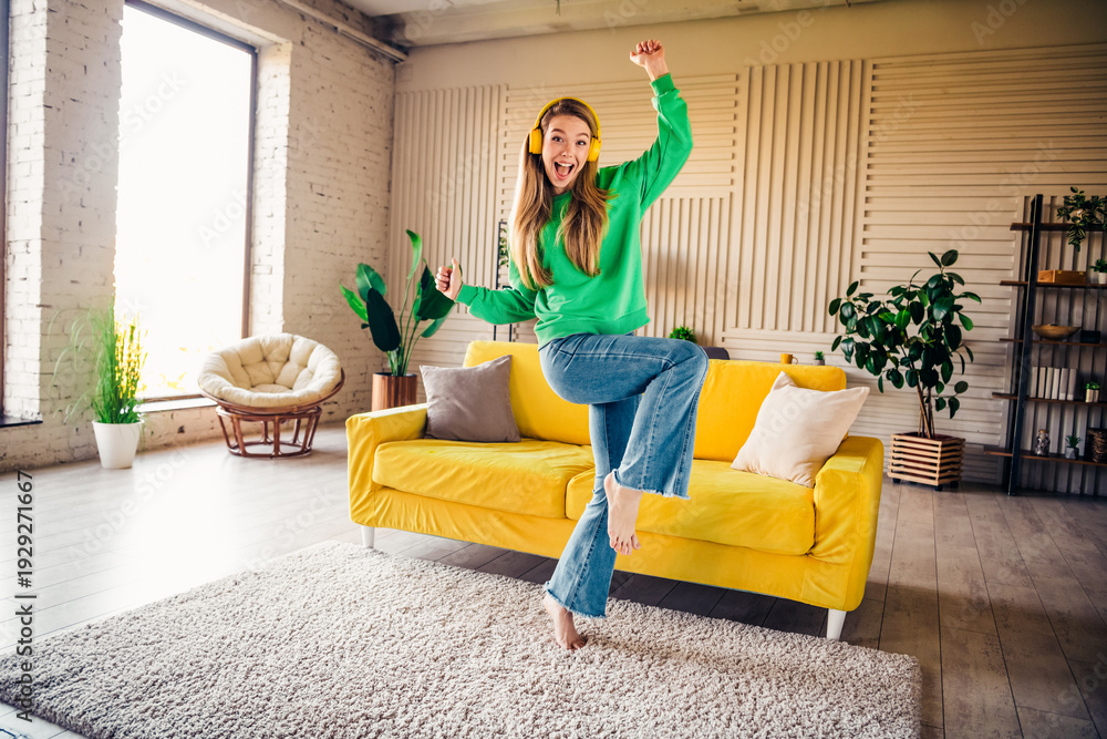 Naklejka premium Young woman in a green sweatshirt cheerfully dancing in a vibrant loft style living room during a sunny day indoors