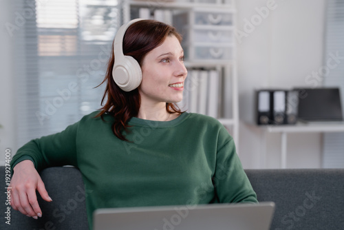 Woman wearing wireless headphones, smiling while enjoying music or a podcast, taking a break from work or studies, creating a peaceful and happy atmosphere in a modern home office setting