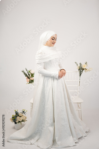 Full length studio portrait of a beautiful bride wearing an elegant lace wedding dress and veil. Minimalist white background with soft floral details creates a timeless and romantic atmosphere. 