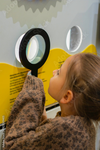 Young girl examines text through a magnifying glass at an interactive museum exhibit with colorful display elements and digital screen