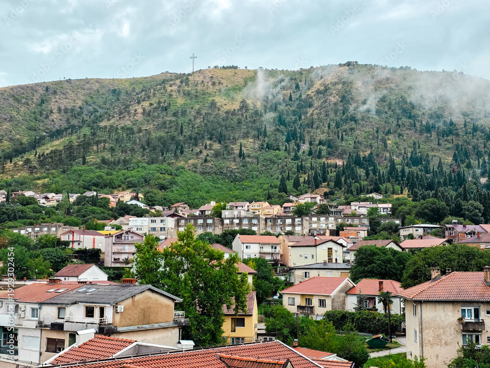 Fototapeta premium Panoramic view of Mostar, Bosnia and Herzegovina with residential district and green hillside. Large cross monument visible on the hill under cloudy sky after rain.