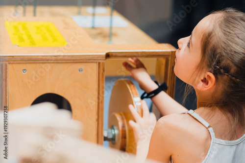 Young girl interacting with a wooden educational exhibit featuring gears and levers inside an interactive museum space designed for childrens learning and exploration