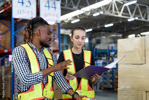 Schilderij op canvas Professional warehouse staff, African American technician and Caucasian manager, analyzing stock list on clipboard during workday inside industrial storage facility, ensuring accurate inventory