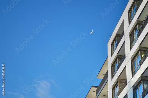 Modern white residential apartment building facade in Warsaw under a bright clear blue sky with a small commercial airplane flying high above in the distance.