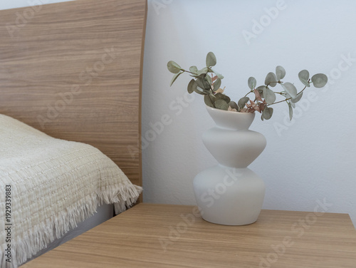 Minimalist bedroom interior with white vase and eucalyptus. Cozy home decor details, white vase on wooden nightstand near bed.