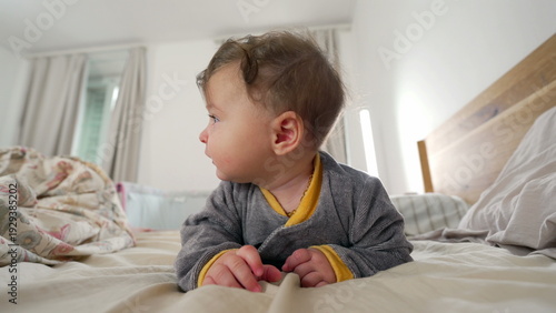 Фотография Baby lying on stomach on bed, smiling softly while bringing hand to mouth, joyfu