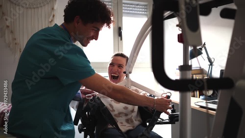 Young male caregiver assisting a laughing disabled woman in a wheelchair, using a patient hoist lift for safe transfer and mobility inside a house, demonstrating compassionate home healthcare
