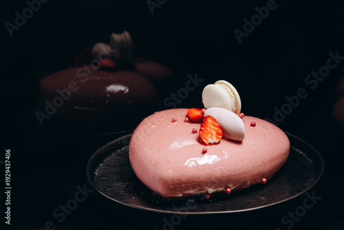 Glossy heart-shaped strawberry mousse cake topped with macarons and fresh berries on black plate