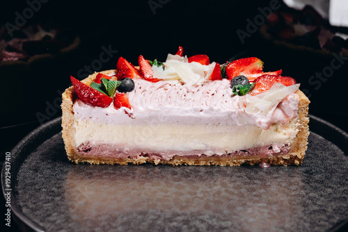 Slice of strawberry cheesecake with whipped cream, fresh berries and coconut shavings on a dark plate