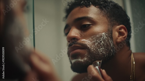 Young man grooming in bathroom with shaving cream on face and razor in hand, focusing on personal care routine