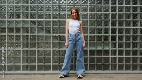 Woman wearing white tank top and blue jeans stands against glass brick wall in a city location