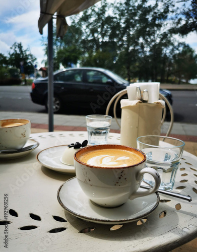 A cup of latte and a small dessert rest on an outdoor café table near glasses of water and street traffic. This image represents casual dining, warm beverages, and relaxed urban moments.