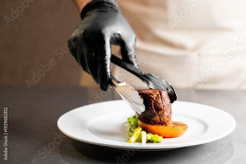 Obraz na płótnie A chef in a black glove uses silver tongs to plate a seared filet mignon over grilled vegetables on a white plate