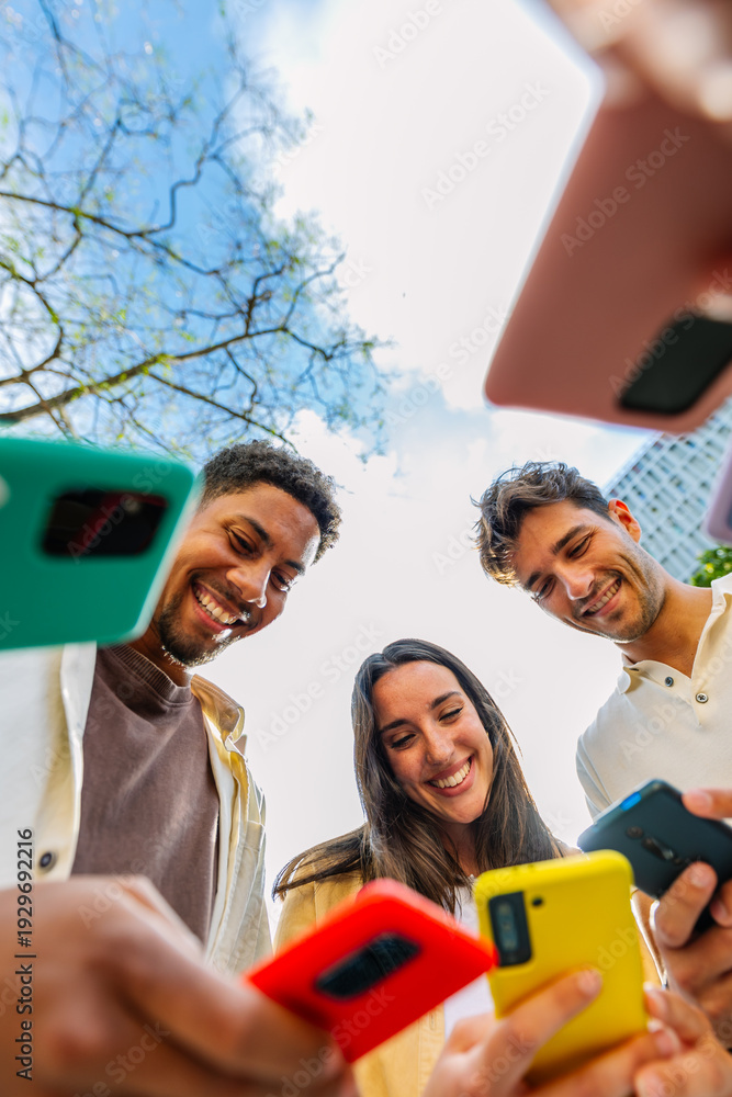 Obraz premium Vertical shot of young group of people using phones devices standing together in circle outdoors. Millennial friends addicted to social media app, betting or playing video game on platform online.