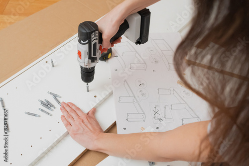 A woman uses a screwdriver to fasten a furniture panel on the floor surrounded by instructions, parts, and tools in home interior