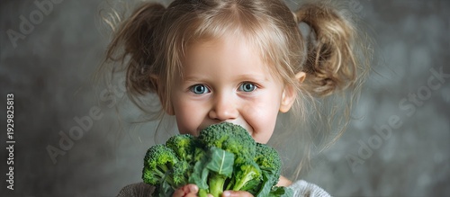 Young Girl with Blonde Hair Blue Eyes Smiling While Holding Fresh Broccoli in Front of Gray Background