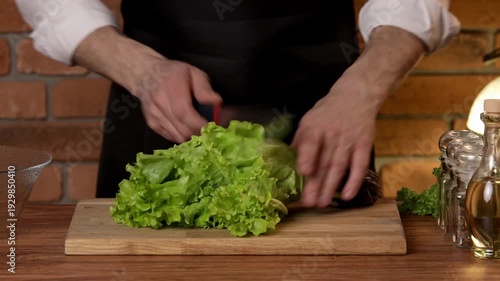 A male chef cuts green salad leaves with a knife on a cutting board.