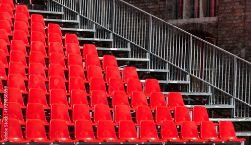 Bright red stadium seats are arranged in rows with a metal railing intersecting diagonally. The image conveys a sense of anticipation and emptiness.