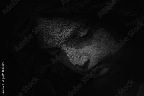 Dark stone statue face emerging from shadows, symbol of sorrow, silence, grief, and spiritual contemplation.