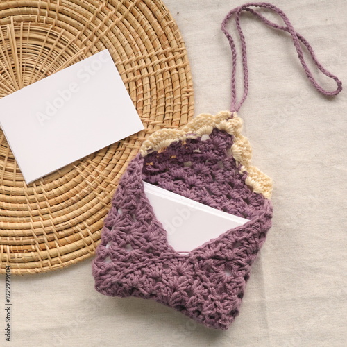 Purple handmade crochet book sleeve with blank white card for copy space on wicker tray and linen background.
