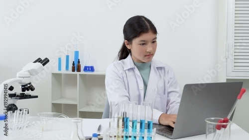 Young Asian women Scientist wearing lab coat writing a report with blue liquid during experiment in modern laboratory with microscope and test tubes rack and laptop
