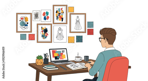 A male graphic designer with glasses sits at a wooden desk working on a laptop and sketchbook in front of a gallery wall of art.