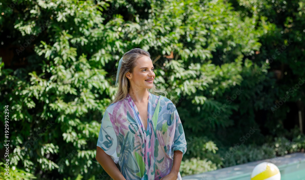 Fototapeta premium Woman standing poolside in sunlit garden wearing pastel floral cover-up near inflatable beach ball