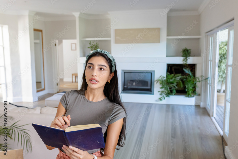 Obraz premium Indian woman standing and holding navy-blue notebook and pen, looking upward in living room