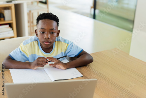 African American child sitting at home study table holding pen writing in notebook near laptop