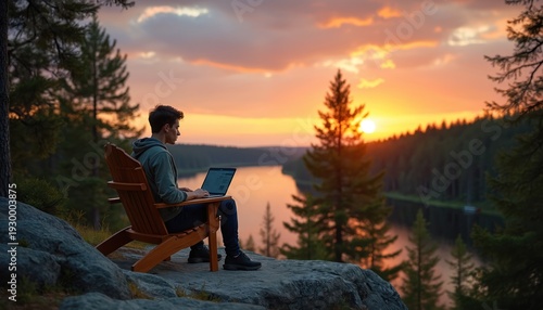 Young man works on laptop in wooden chair on rock near lake. Forest landscape at sunset. Person uses computer outdoors in nature. Peaceful remote lifestyle scene.