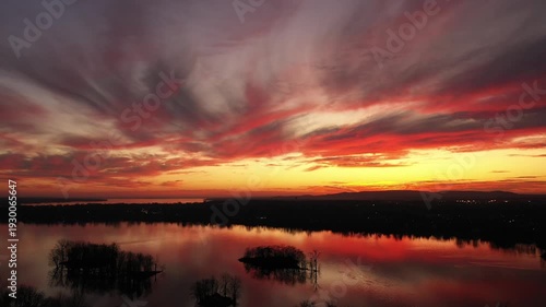High altitude drone shot, panning gently from right to left, at the end of sunset, with dramatic colors of orange, gold, and turquoise contrasting with the black trees in the calm river