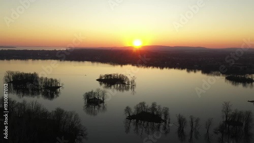 Drone shot, descending gently over calm water, behind the silhouette of a bare tree, during a sunset in a cloudless sky