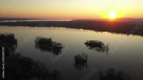 Drone shot, gently rotating to the left over calm water, dotted with small islands, showing a relaxing landscape during a sunset in a cloudless sky, behind mountains