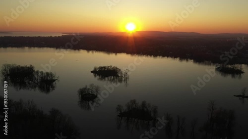 Drone shot, moving gently from right to left over calm water, dotted with small islands, showing a sunset in a cloudless sky, behind mountains