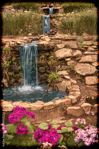 A serene garden features a cascading waterfall surrounded by stone walls and vibrant flowers. The natural setting creates a peaceful ambiance.