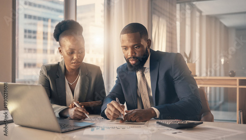 Professional african american business partners working on laptop together in modern office environment with financial documents and calculator discussing company growth and strategy during day