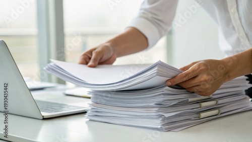Person organizing a large stack of documents on a desk beside a laptop in a bright office setting.