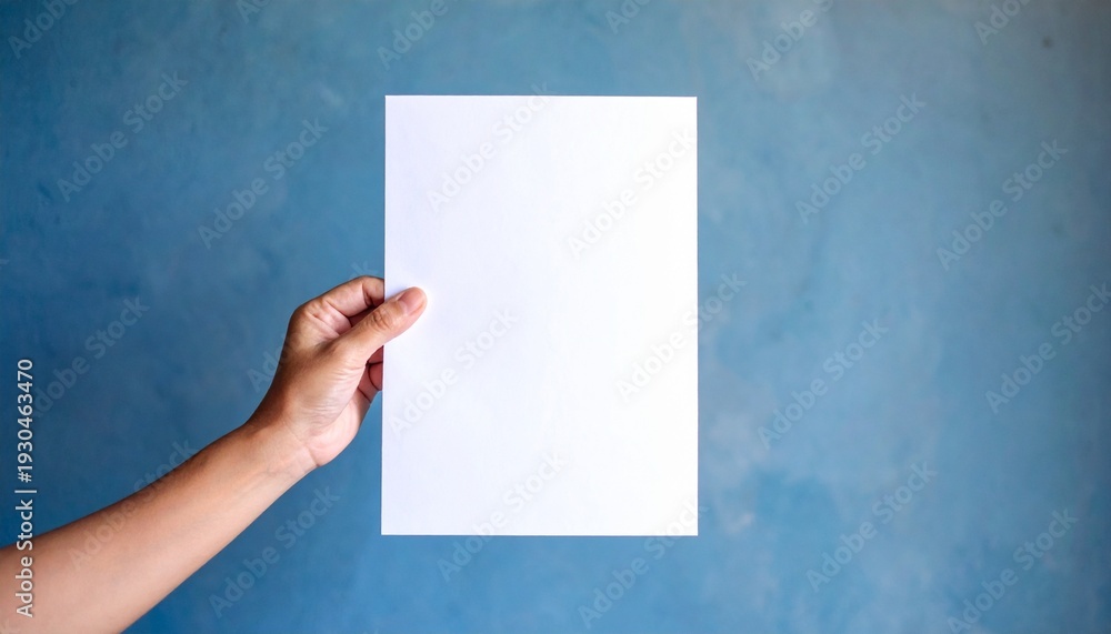Obraz premium Hand holding a blank white paper against a textured blue wall for design mockups and presentations.