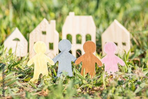 Colorful wooden people figures holding hands in front of wooden house models on green grass, community, real estate concept. Real estate agency. Renewable and energy-efficient
