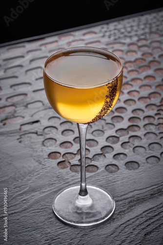Fotografija An elegant amber-colored cocktail served in a sleek long-stemmed coupe glass, featuring a delicate dark spice rim along one side, presented on a dark textured surface